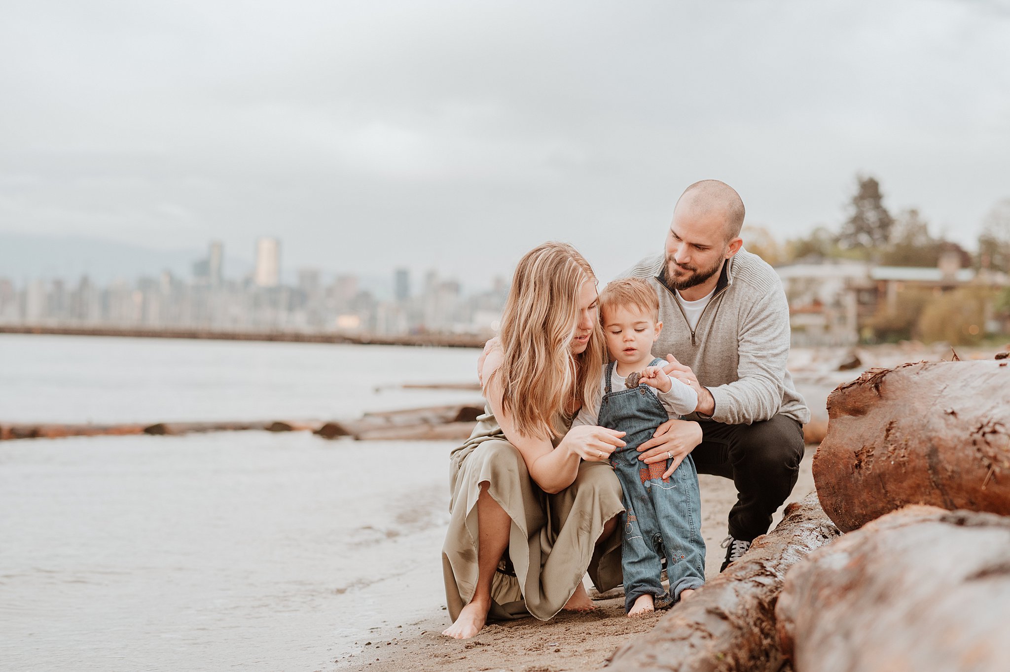 A mom and dad explore rocks on a beach at sunset with their toddler son in overalls after visiting chorus and clouds