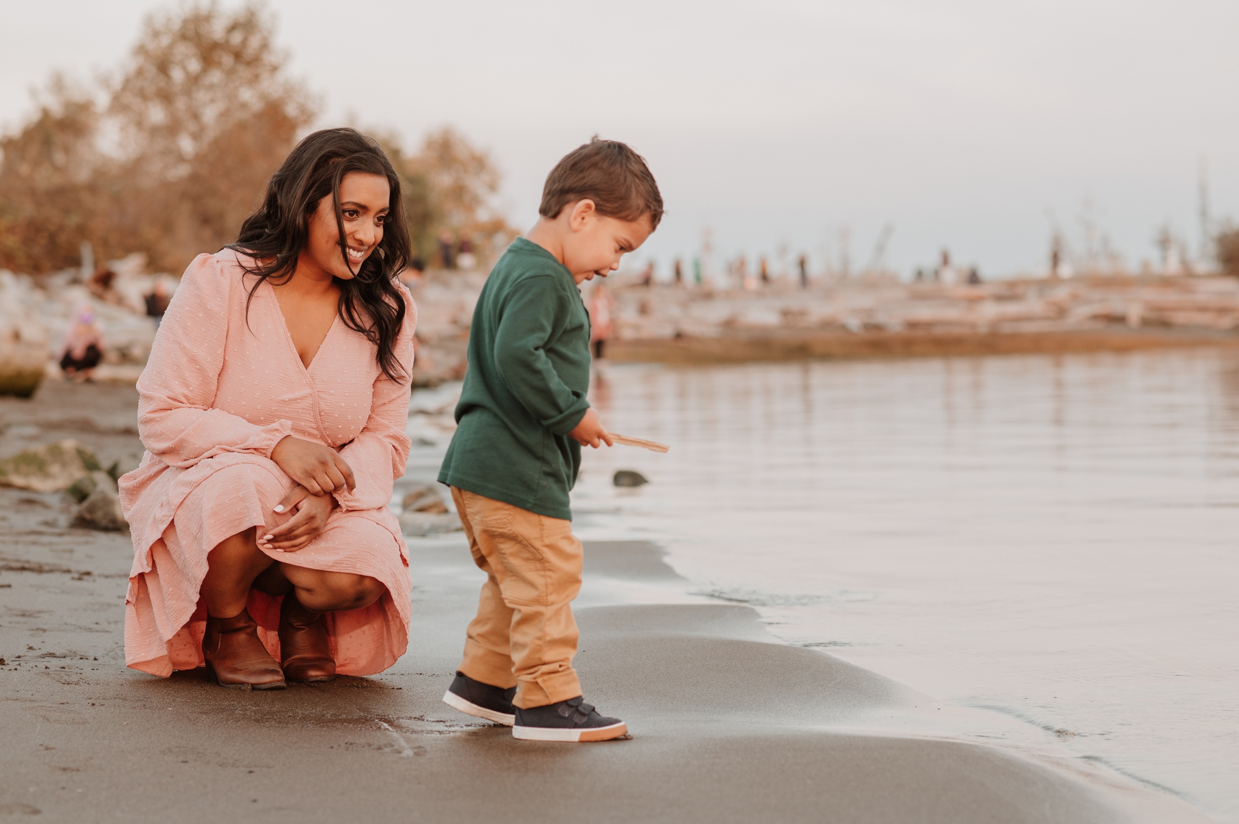 A toddler boy in a green shirt explores the water's edge with a stick while being watched by mom