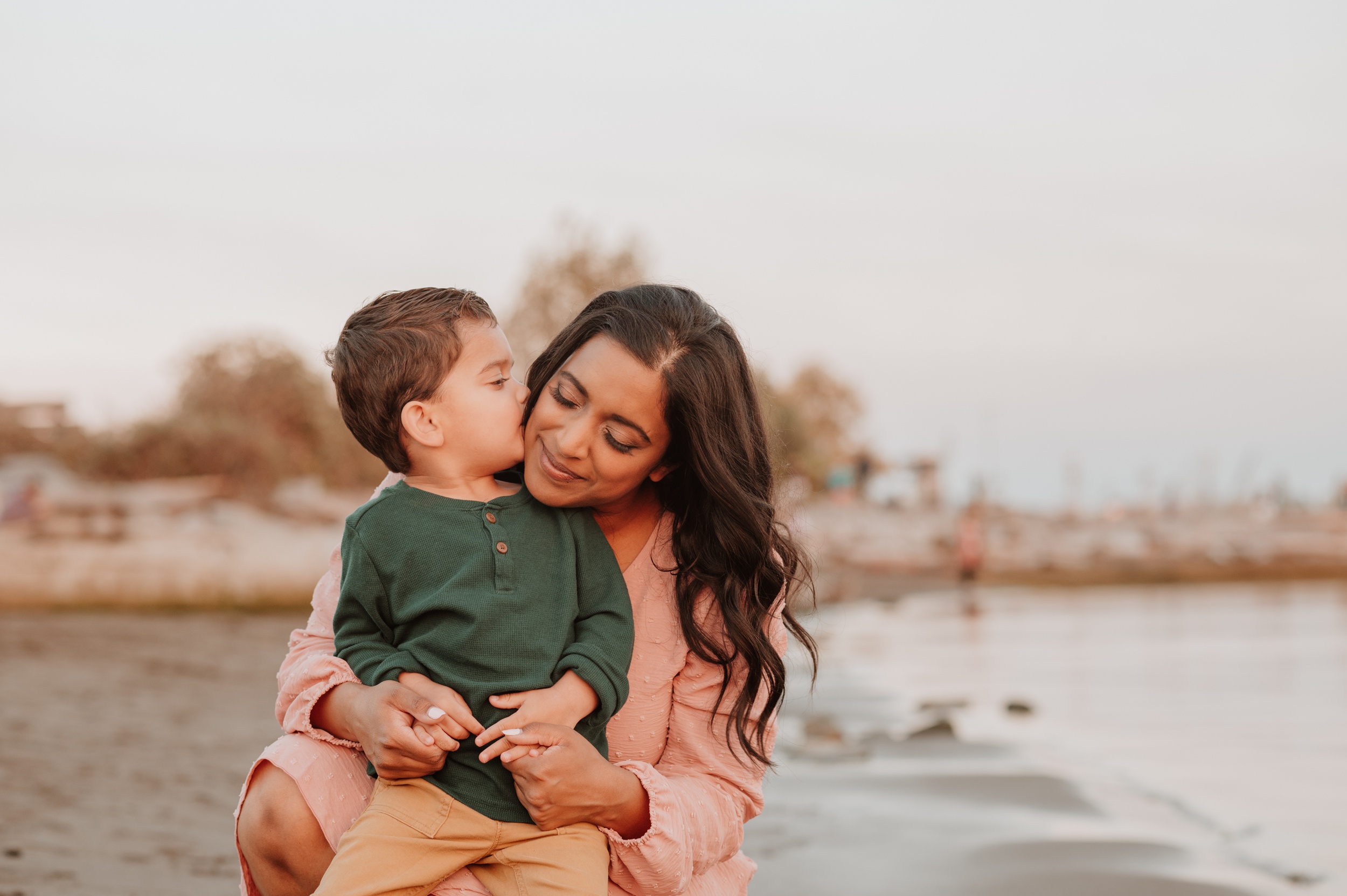 A happy mom snuggles and is kissed by her toddler son on a beach at sunset before visiting JelloLand