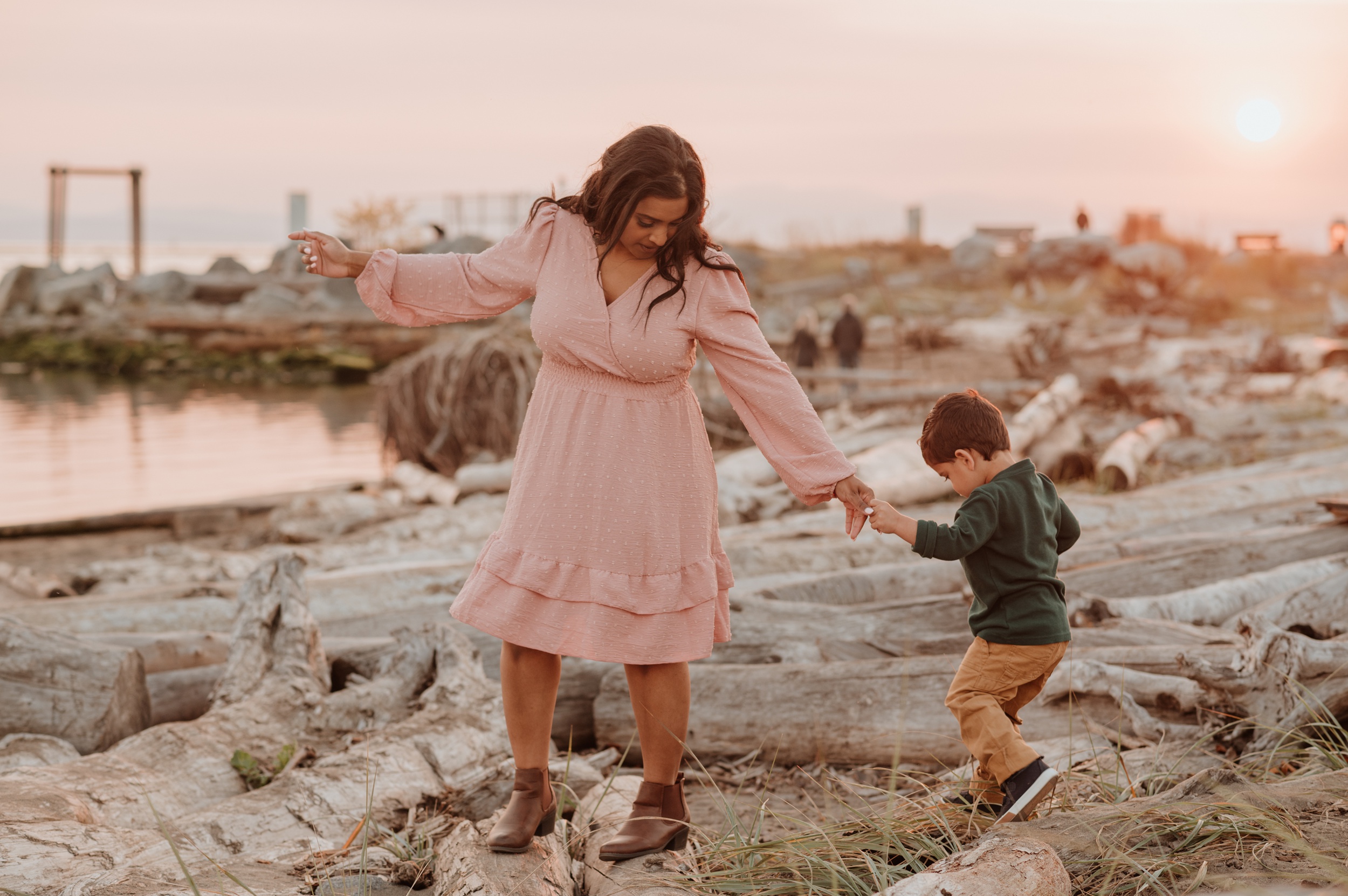 A mom in a pink dress helps her toddler walk on driftwood on a beach at sunset before visiting JelloLand