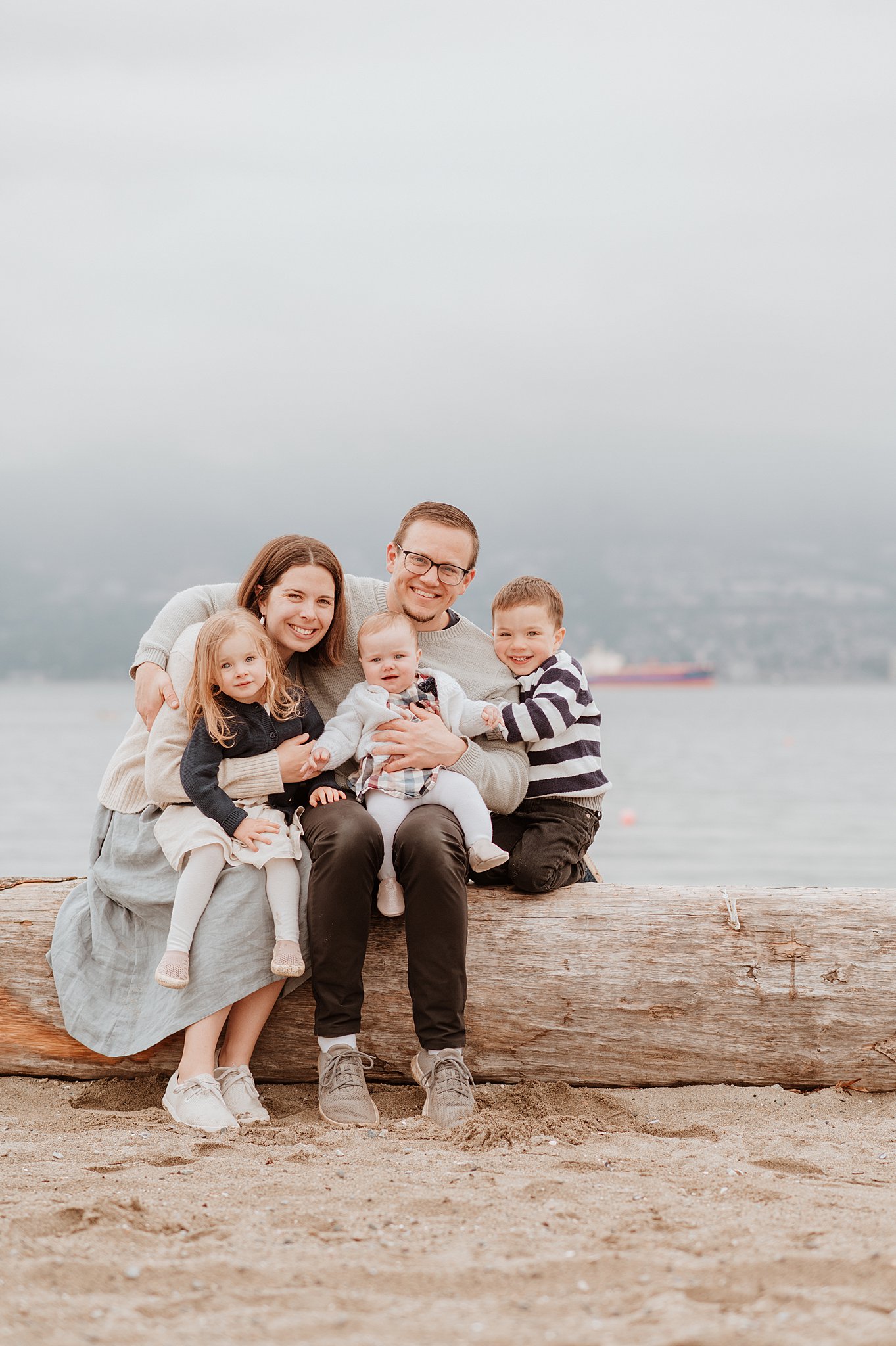 A happy mom and dad sit on a beach driftwood log hugging their three toddlers