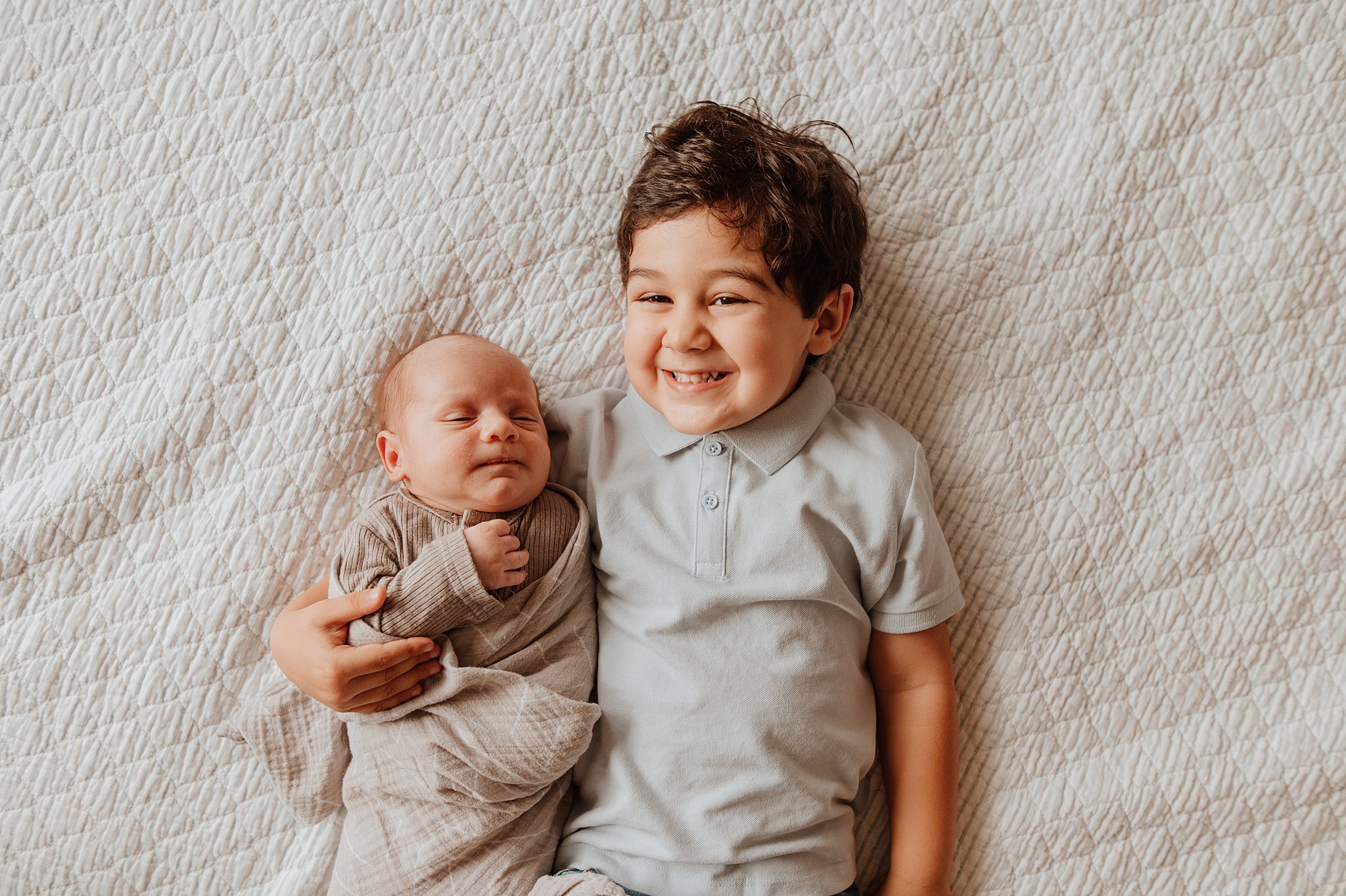 A smiling toddler boy lays on a bed cuddling his newborn baby sibling