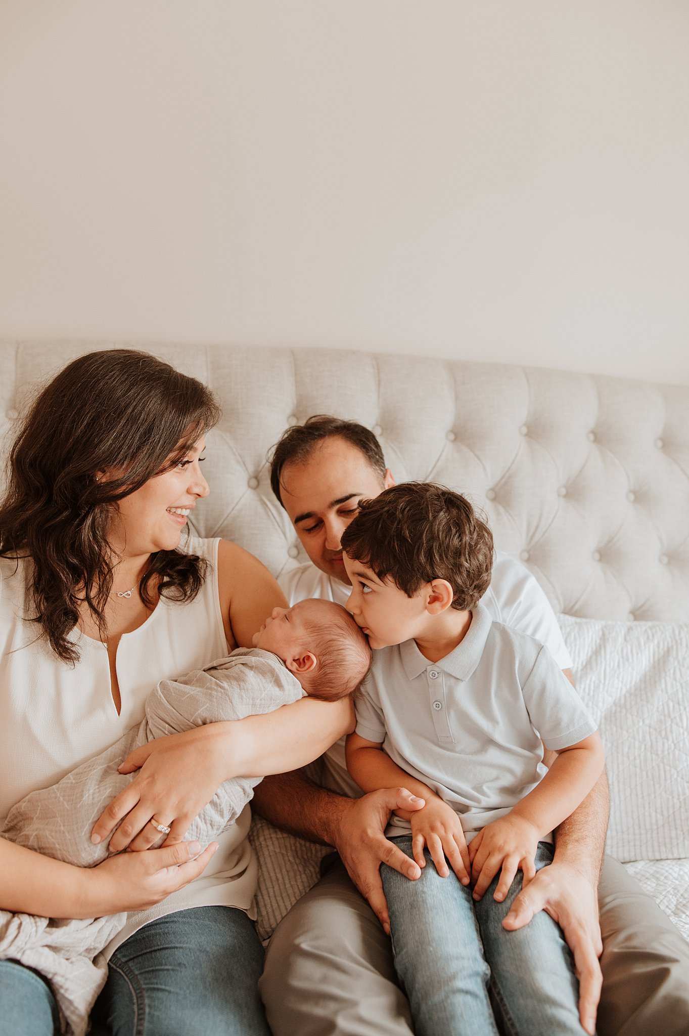 A toddler boy sits in dad's lap on a bed and kisses his newborn sibling's head after visiting knotty toys