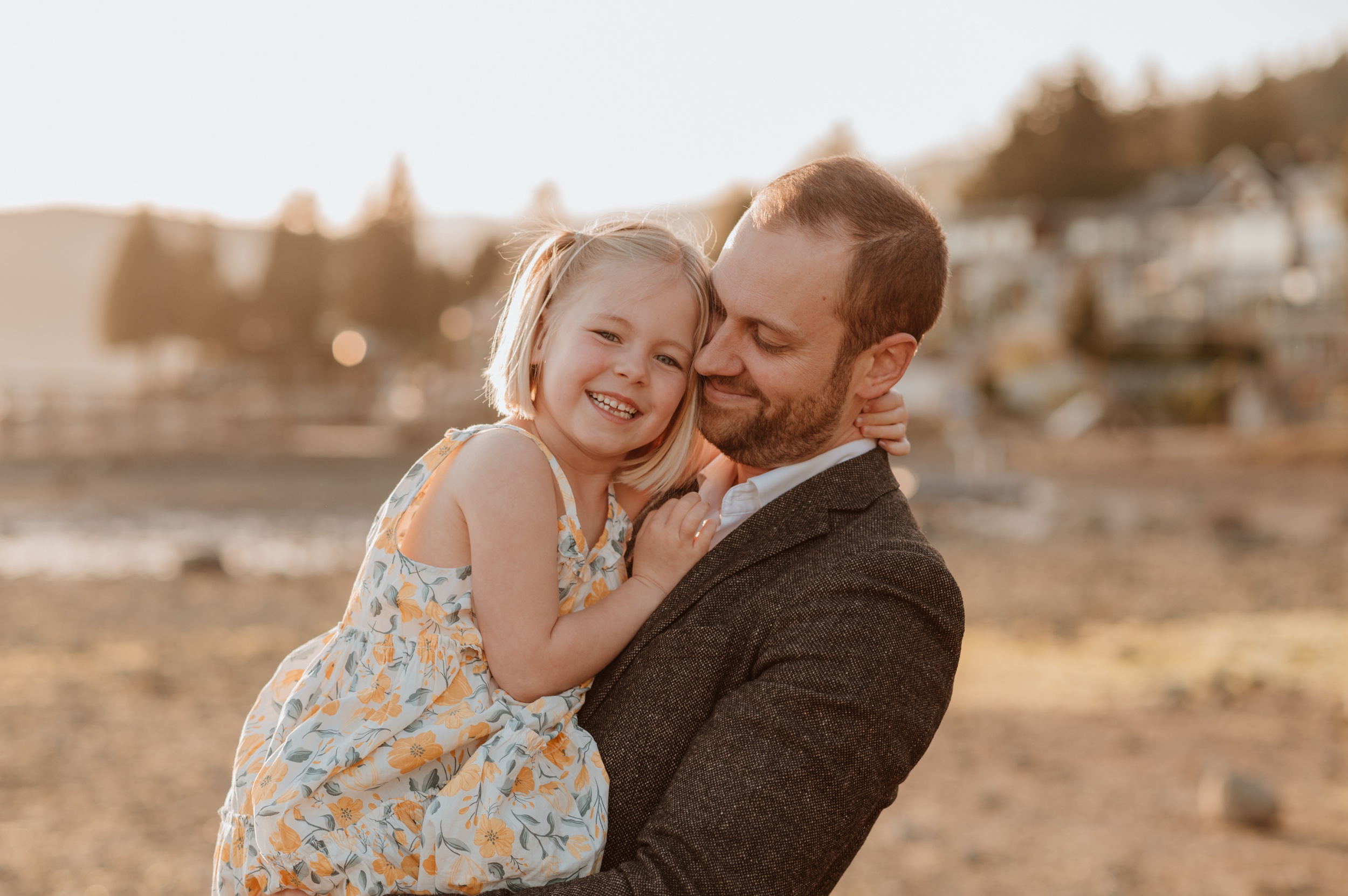 A young girl in a yellow floral dress hugs her dad and smiles while sitting in his arms on a beach after visiting playscape cafe