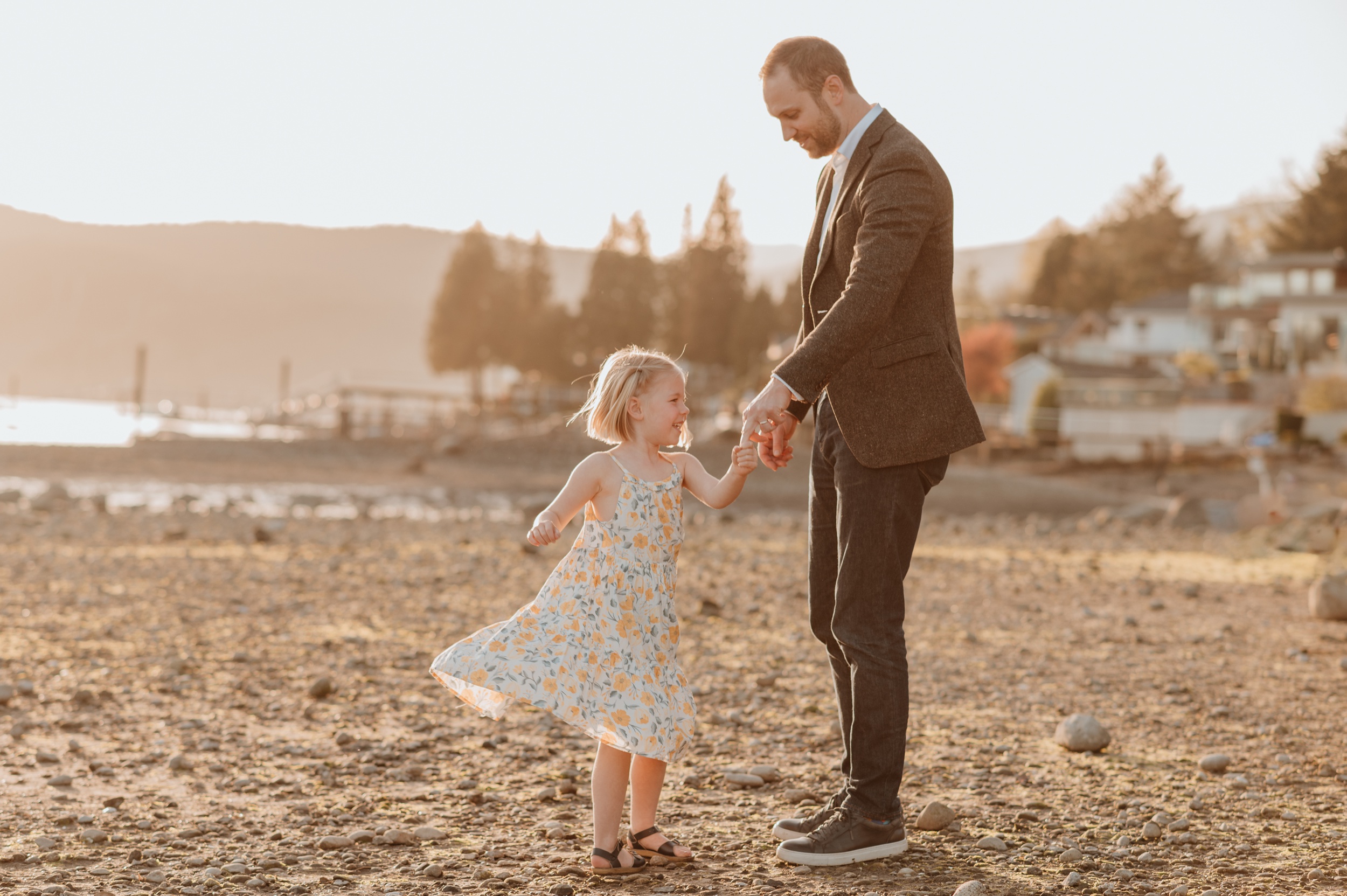 A young girl in a floral dress dances with dad on a beach at sunset after visiting playscape cafe