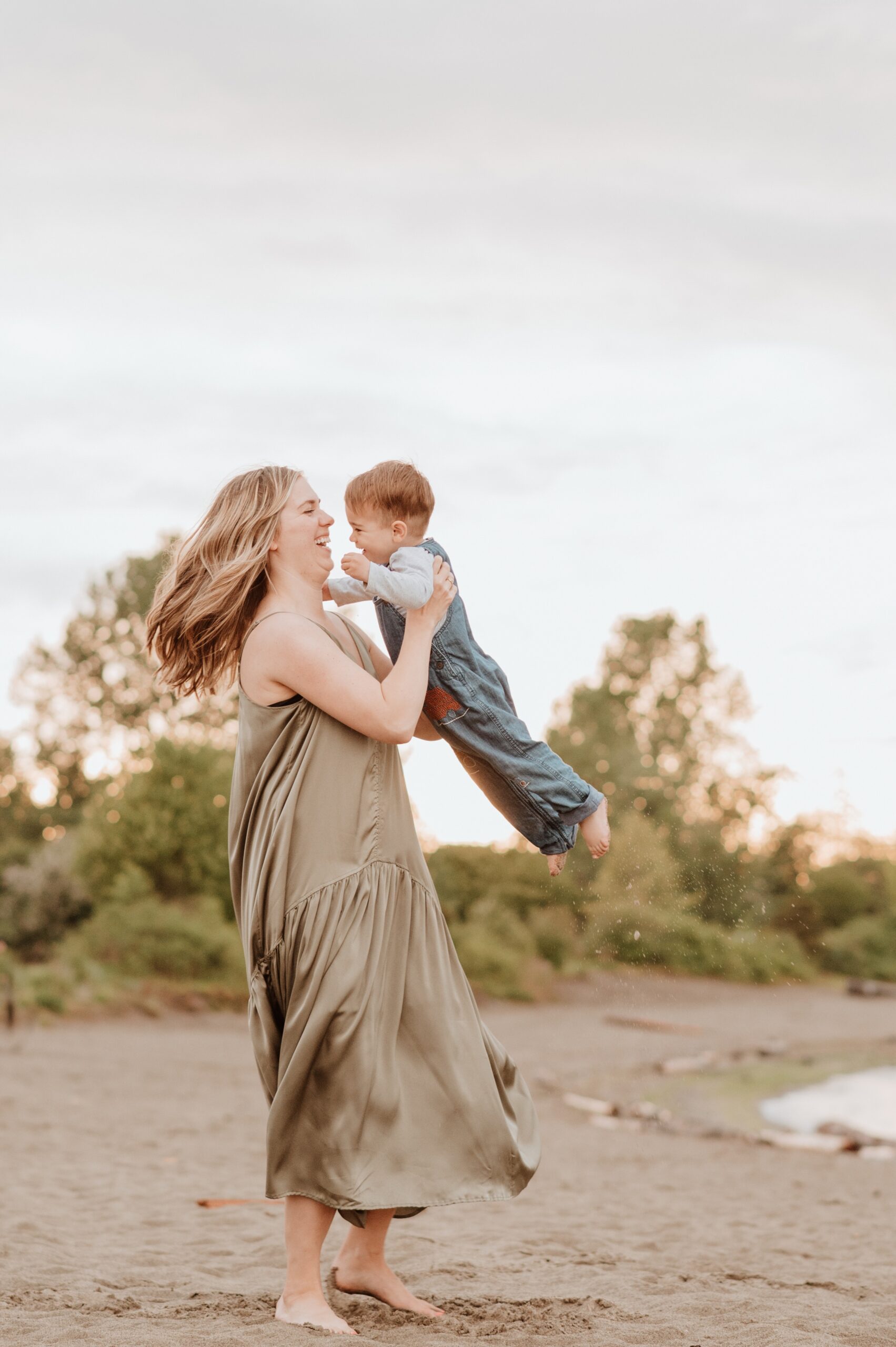 A mom spins and plays with her giggling toddler son on a beach in overalls and a green dress