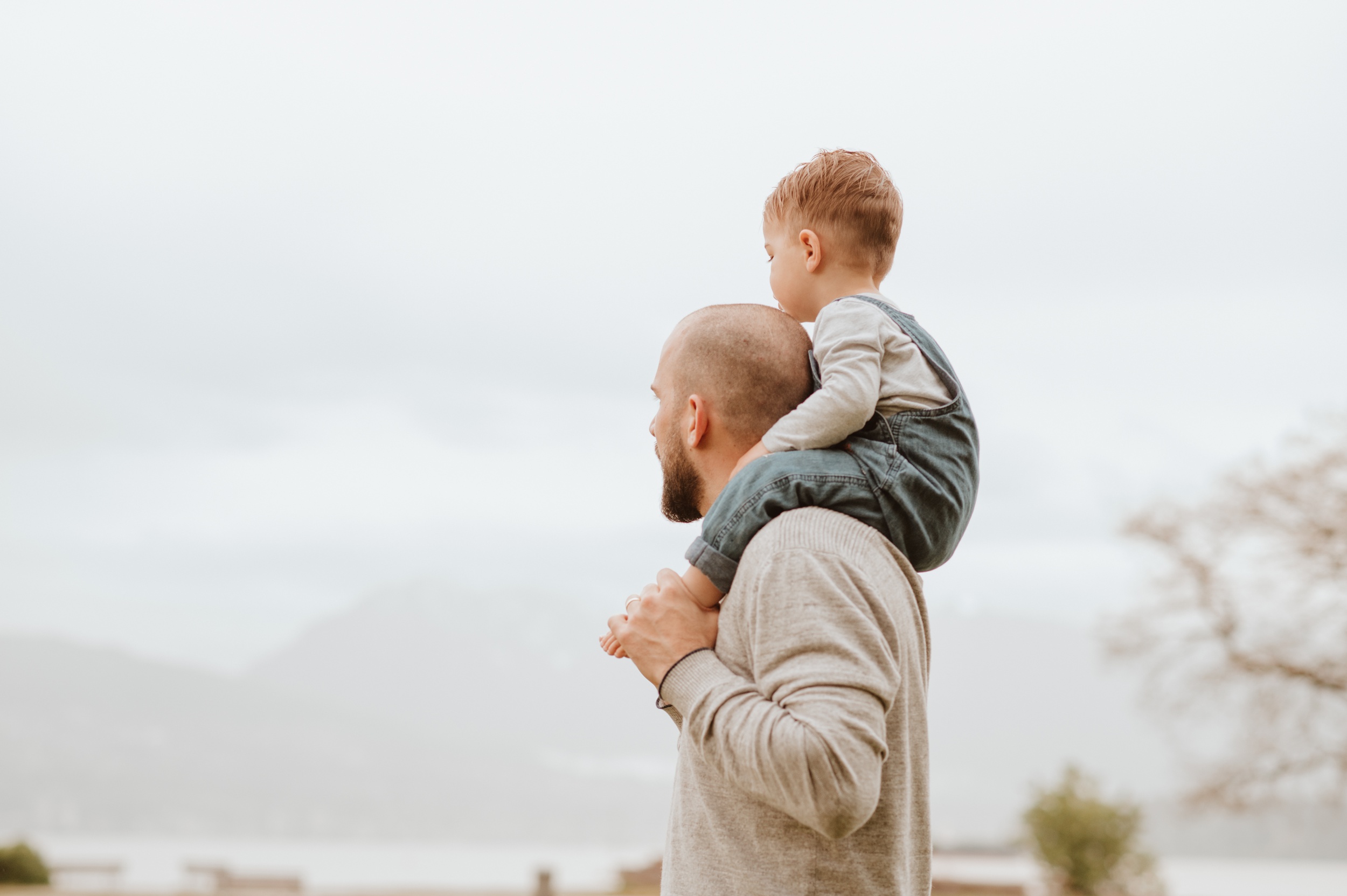 A toddler in overalls sits on dad's shoulders exploring a beach after visiting hip baby vancouver