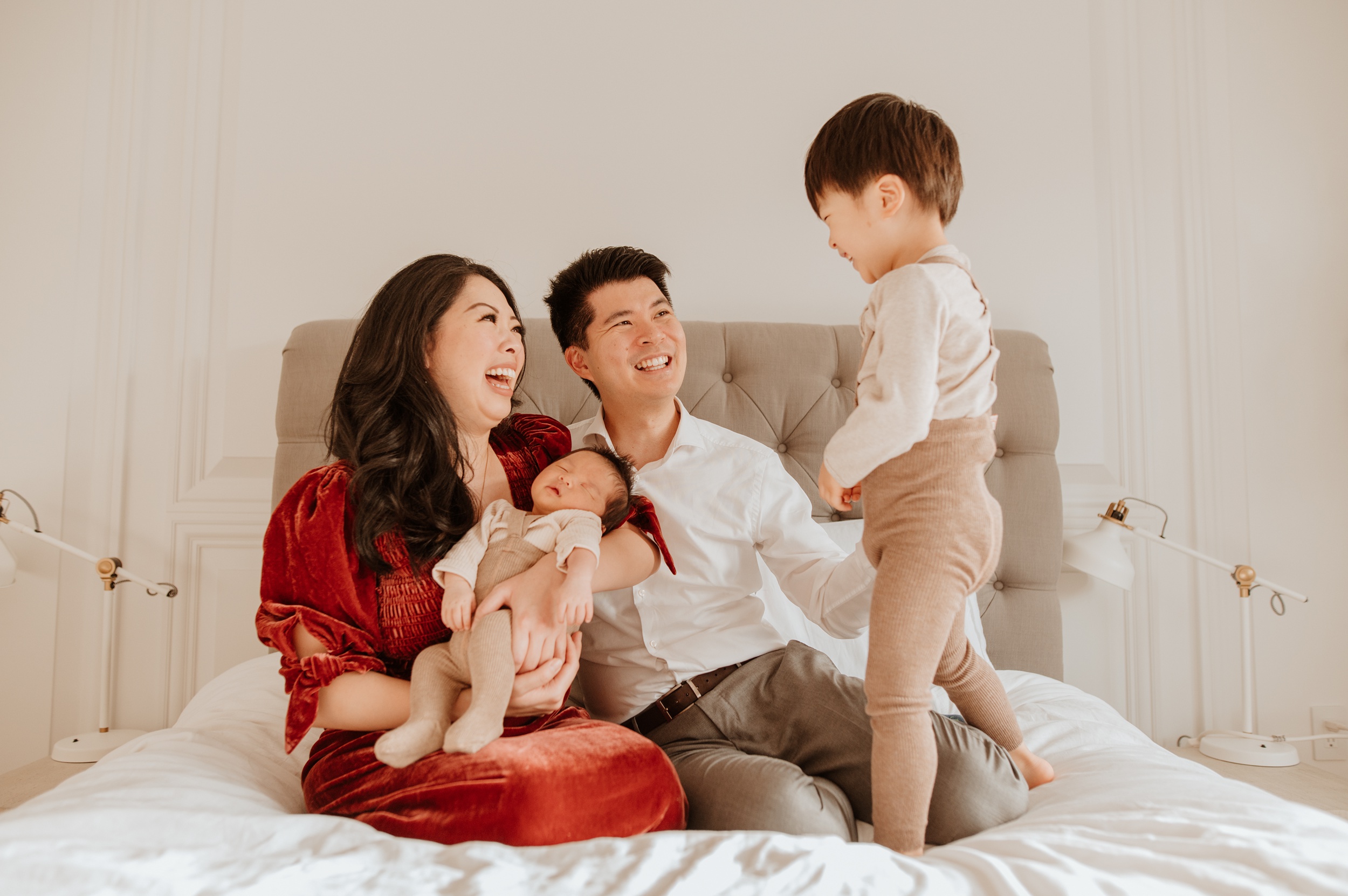 A happy toddler giggles while standing on a bed over mom, dad and sleeping newborn sibling after visiting indoor playgrounds in langley