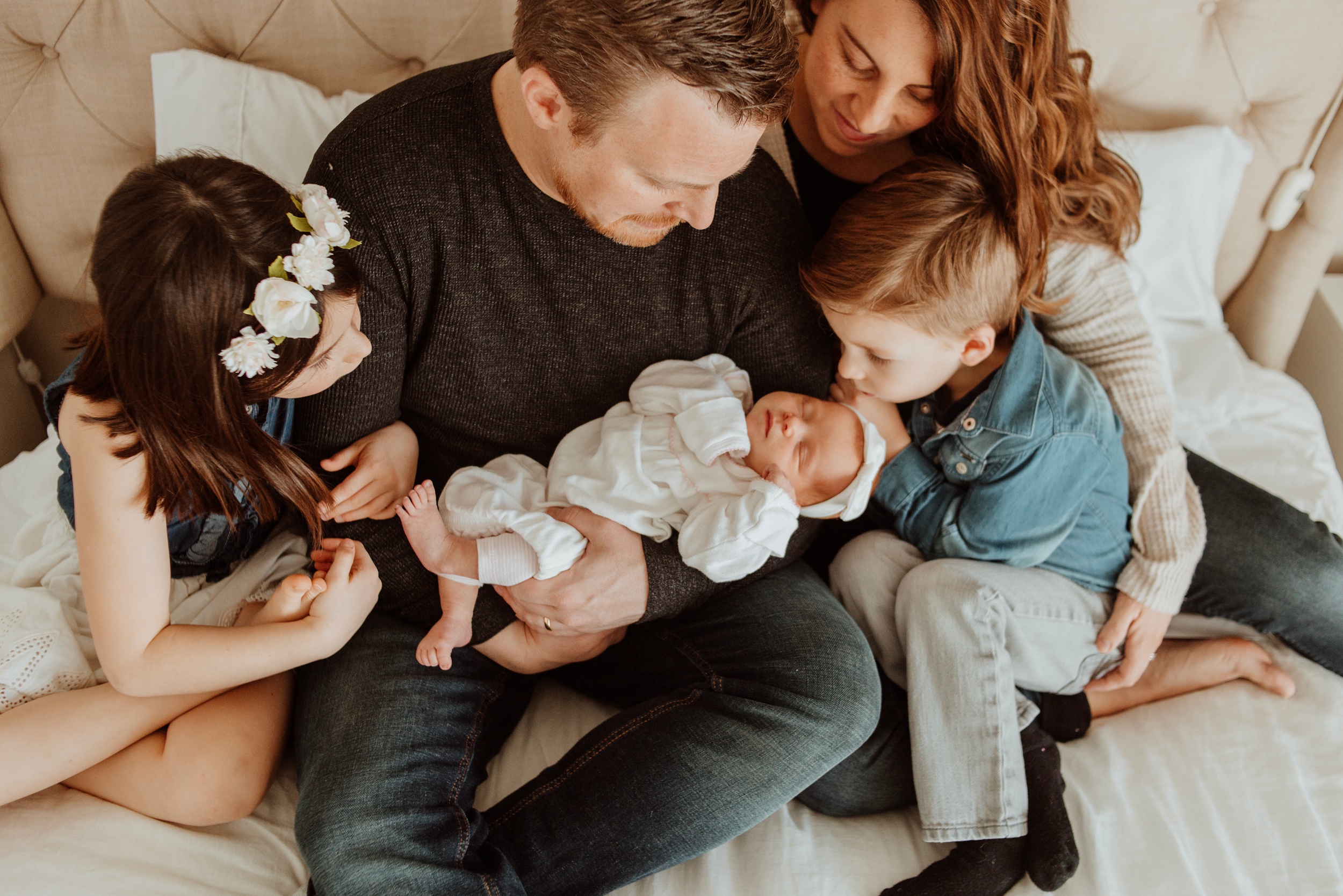 A newborn sleeps in dad's lap on a bed with older siblings and mom admiring it before visiting indoor playgrounds in langley