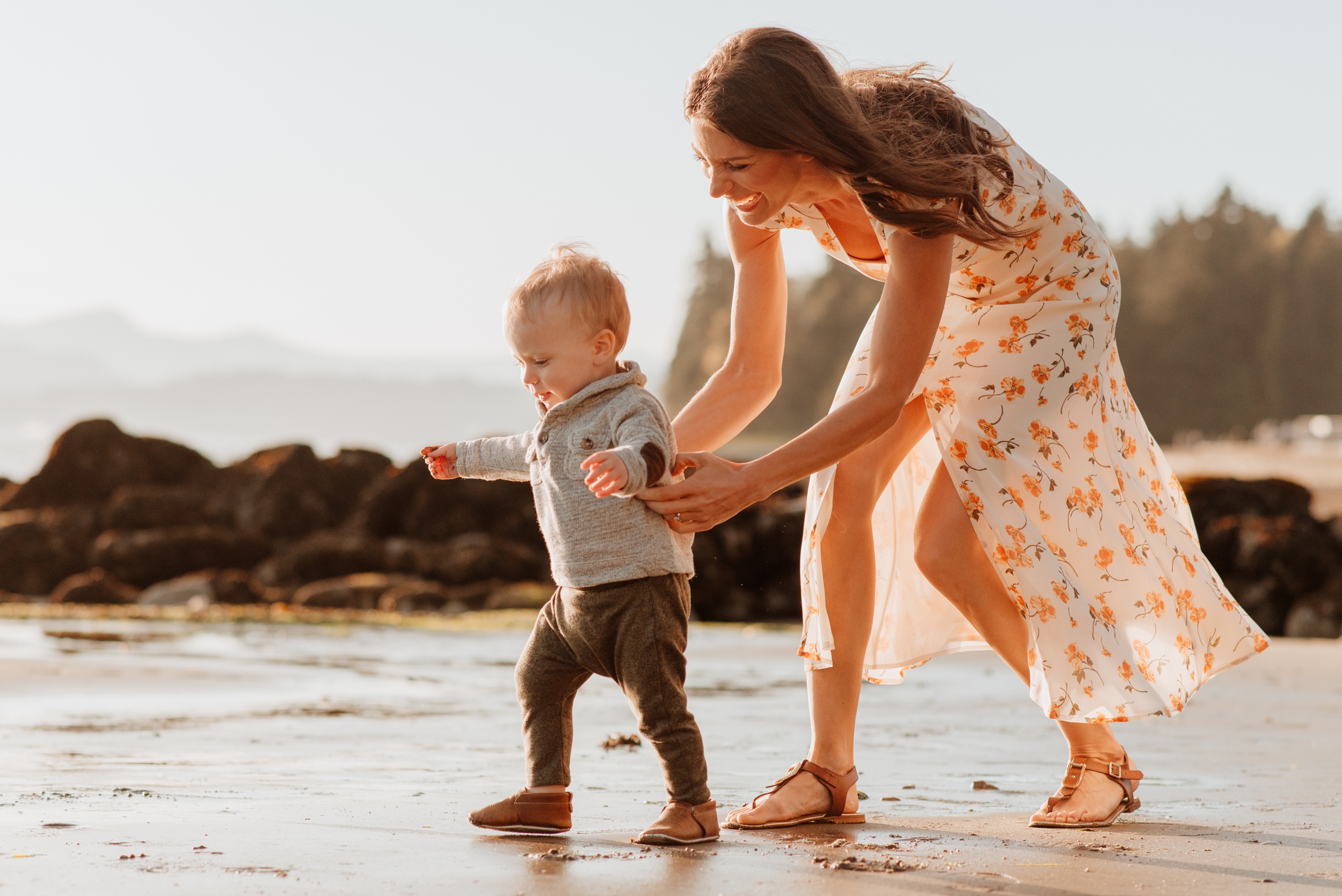 A mom chases her toddler on a beach at sunset