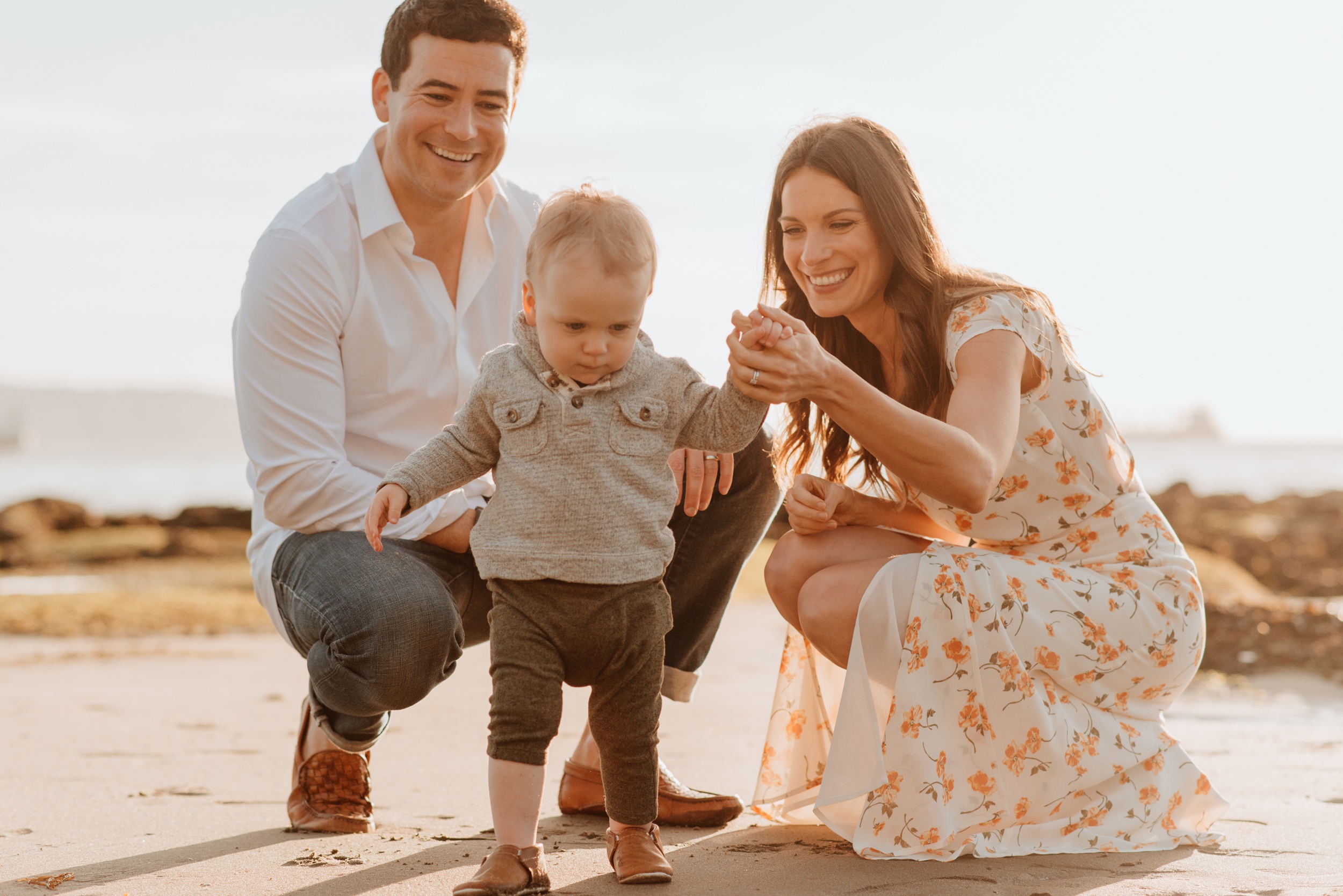 AA toddler walks on a beach in a sweater with help from mom in a floral dress at sunset after visiting lilly and frank