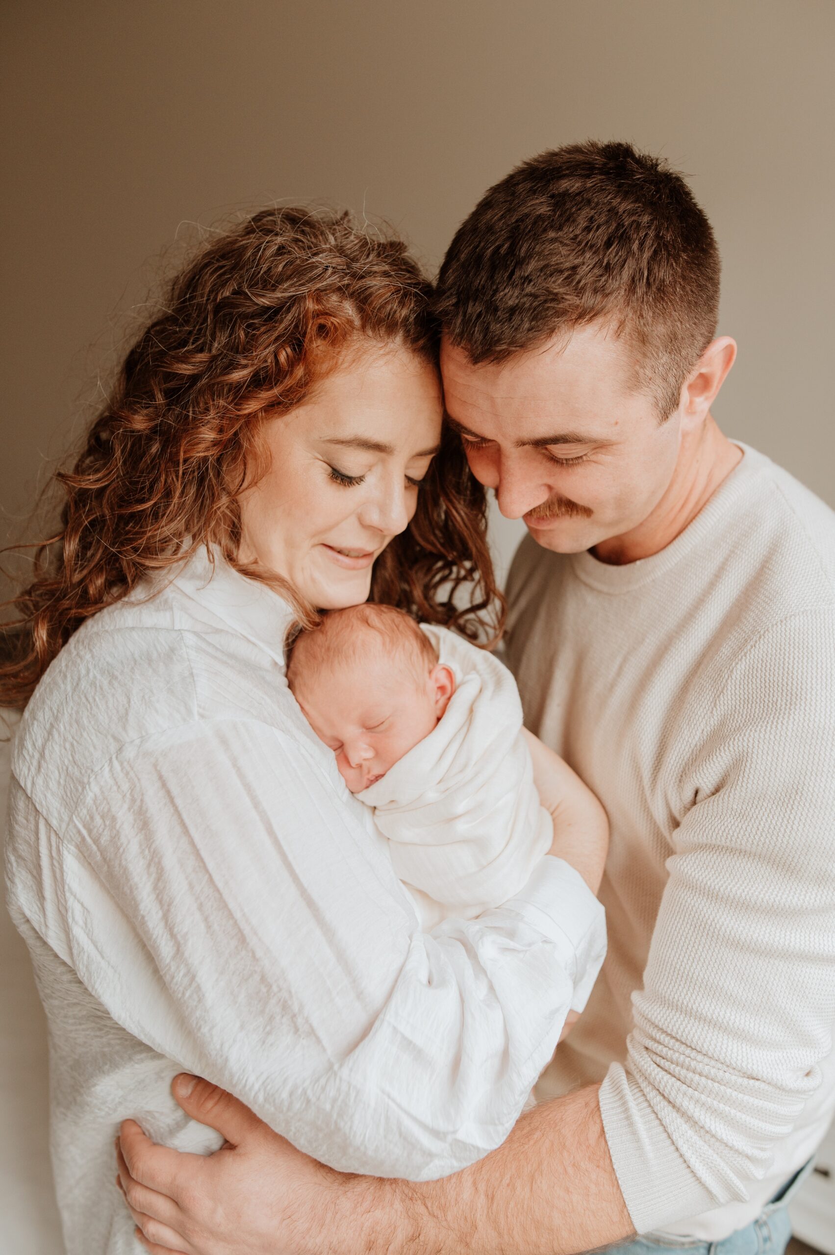 Happy new parents in white snuggles their sleeping newborn in a white swaddle against mom's chest