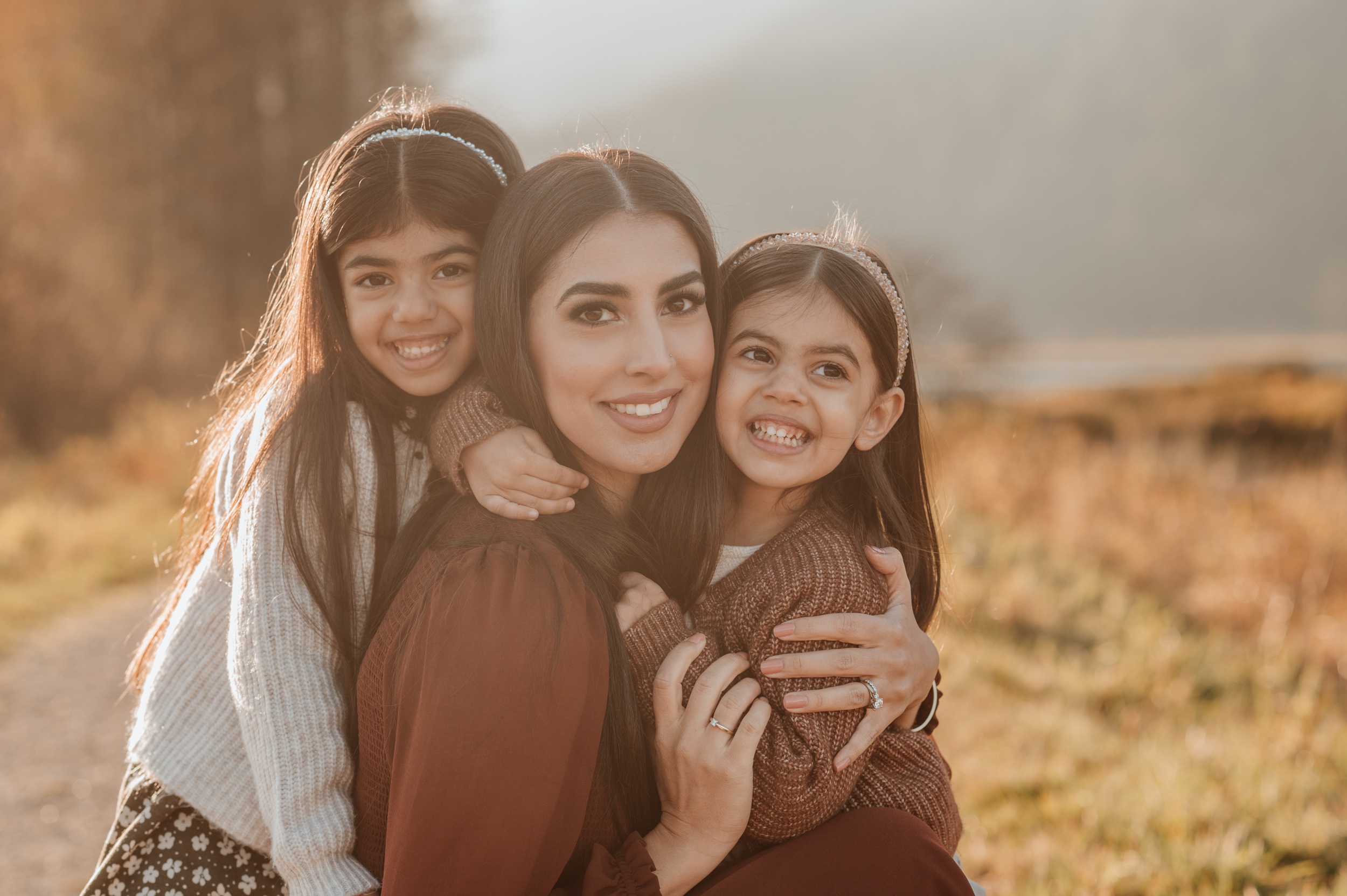 Toddler sisters hug and smile with their mother in a lakeside trail