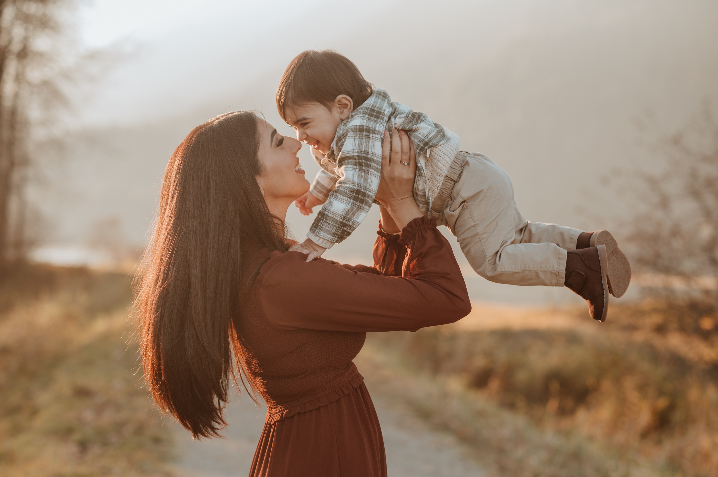 A giggling toddler boy is lifted by happy mom in a brown dress in a park trail at sunset after visiting pediatric dentists in Langley, BC