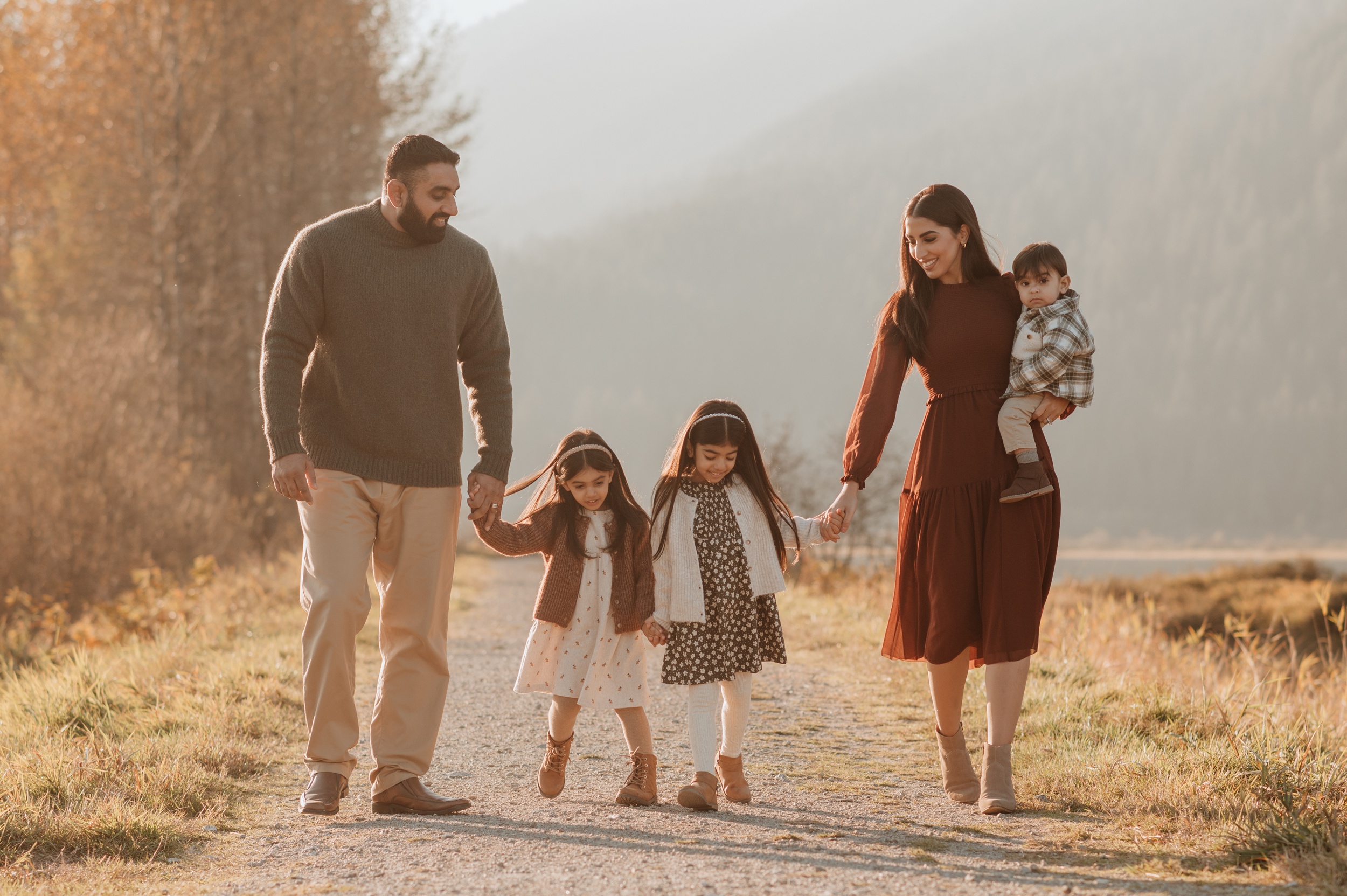 Mom and dad walk their toddler daughters in dresses and hold their infant boy while walking along a lake trail at sunset after visiting pediatric dentists in Langley, BC
