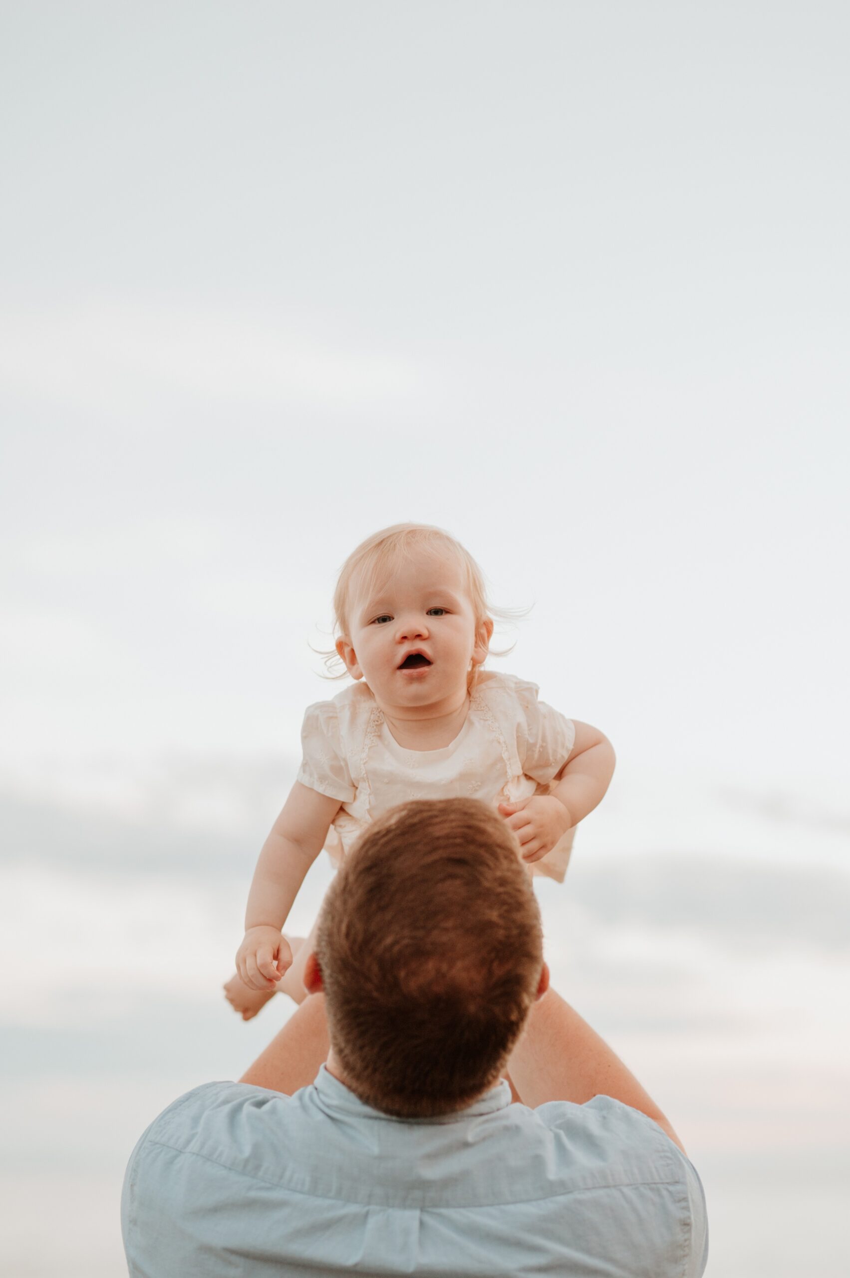A baby girl in a white dress is lifted by dad above his head at sunset after visiting pediatricians in Langley, BC