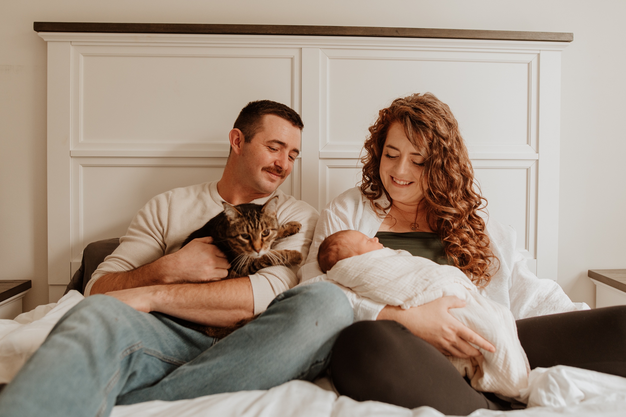 Happy new parents snuggle on a bed with their newborn baby in mom's lap and cat in dad's after she used prenatal massage in Langley, BC