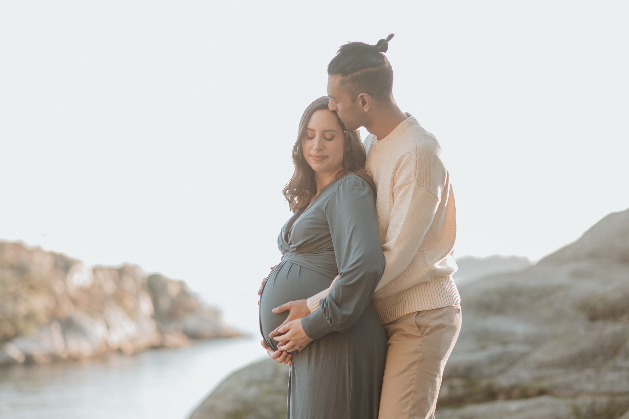A mom to be holds her bump with her husband as they snuggle on a waterfront cliffside at sunset