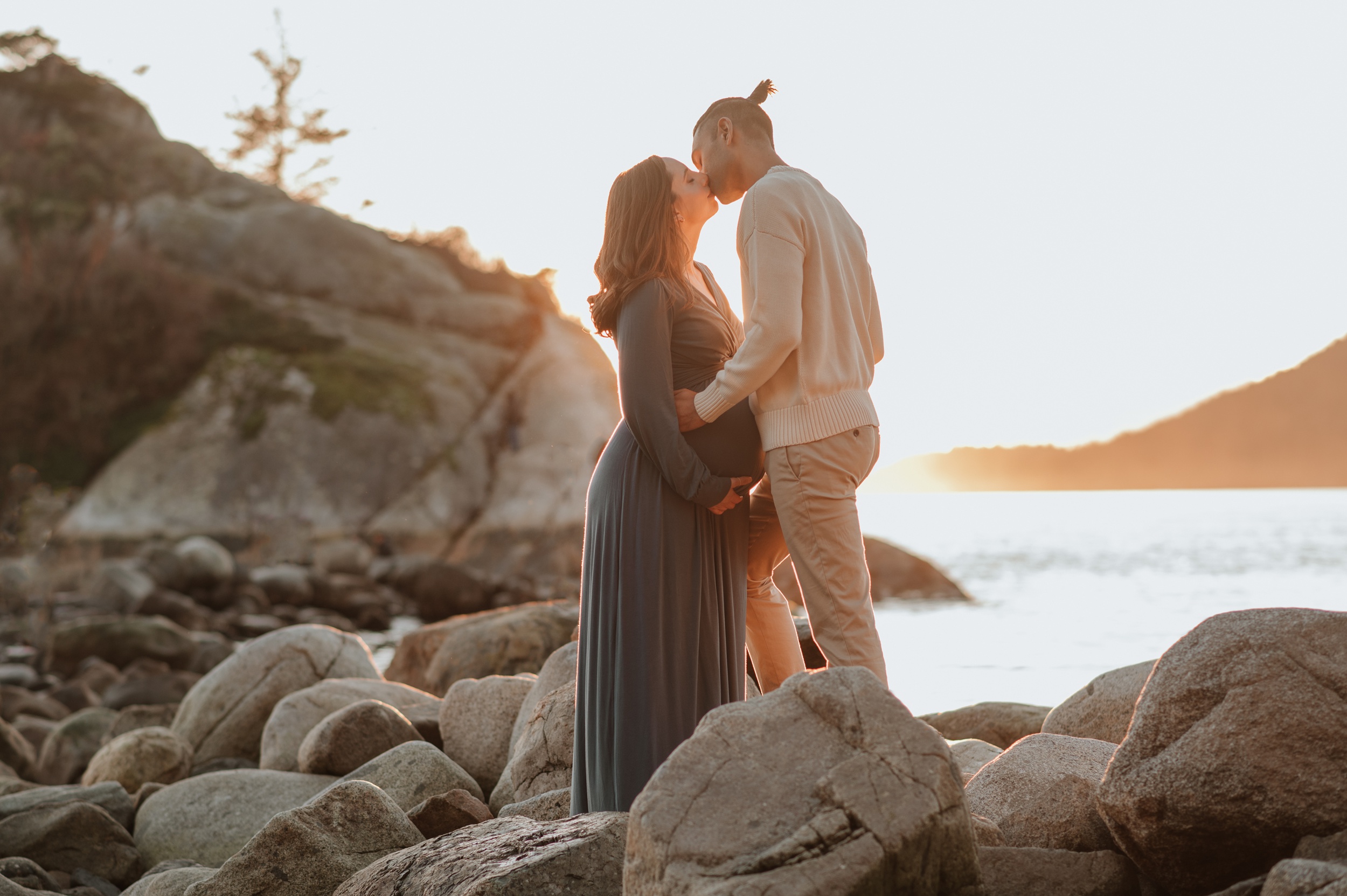 An expecting couple enjoys a romantic kiss on lakeside rocks at sunset after some prenatal yoga in Langley, BC